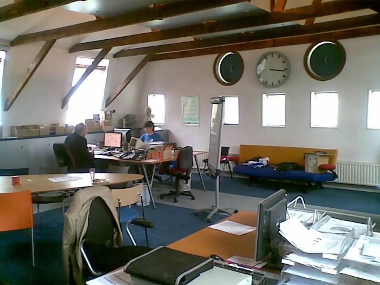 The AnnieS office interior: an open room under exposed wooden roof beams, with two people working at desks, a bank of wall clocks, a flip chart, and stacked paperwork.