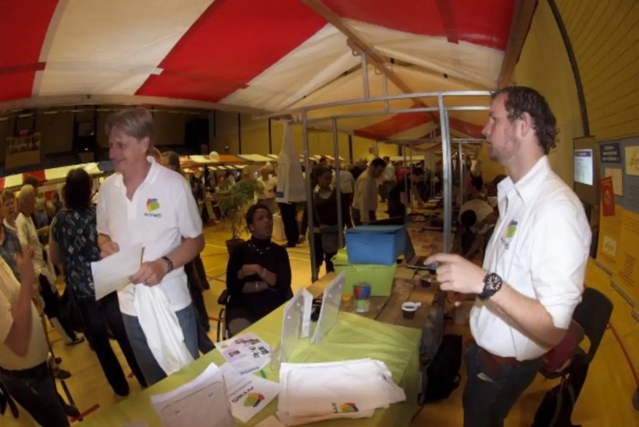 Two AnnieS staff working a stand under a red-and-white striped tent in a sports hall at Werelddovendag 2009: Raymond Kruyer in a branded polo shirt on the left, and Robert McConnell in a white shirt on the right, with flyers and folded AnnieS t-shirts on a green table and visitors crowded around.