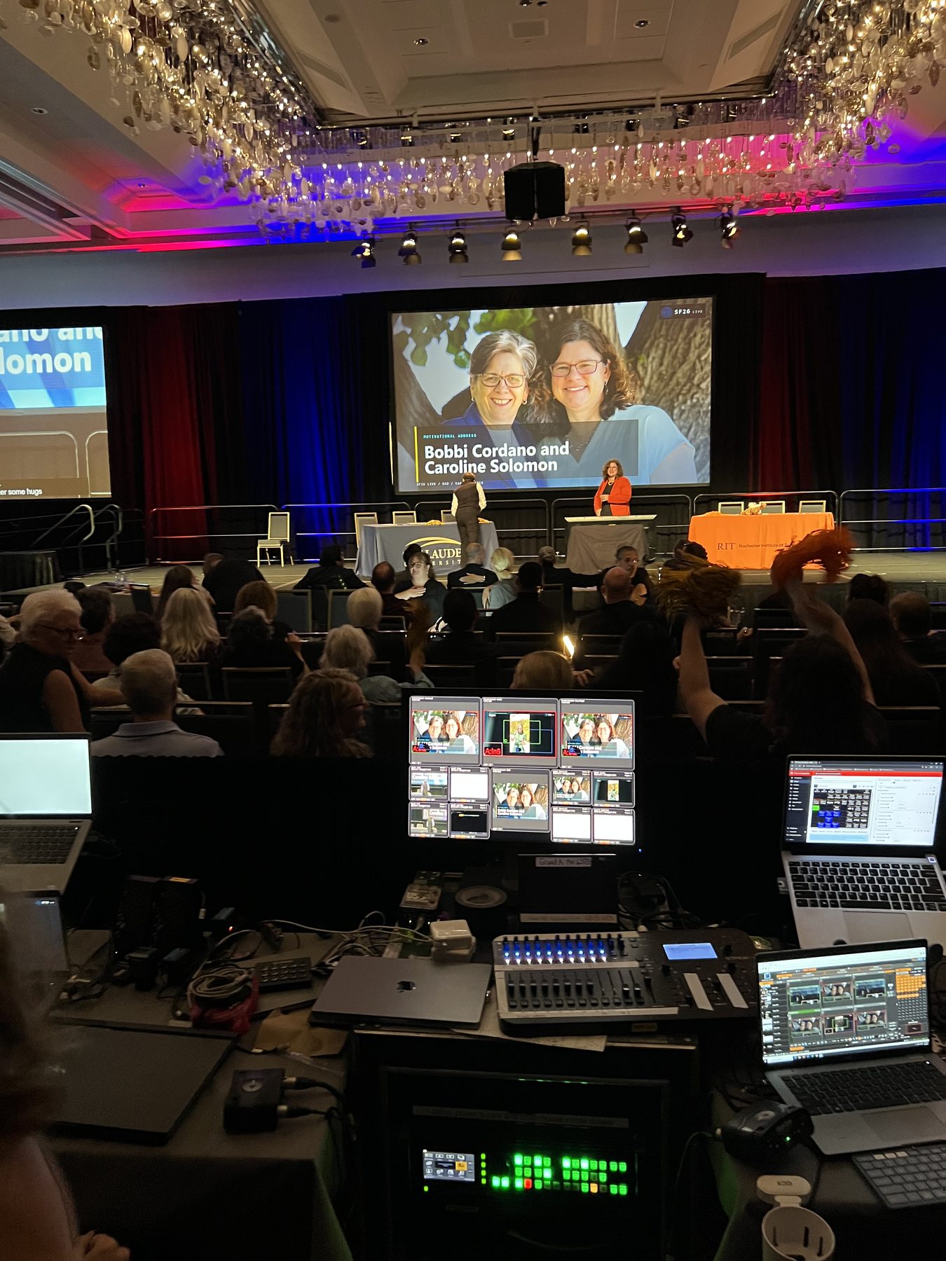 A dimly lit production booth at the back of the Grand Ballroom: multiview monitors, a video switcher and control surface, laptops, and the main stage screen ahead showing a speaker slate.