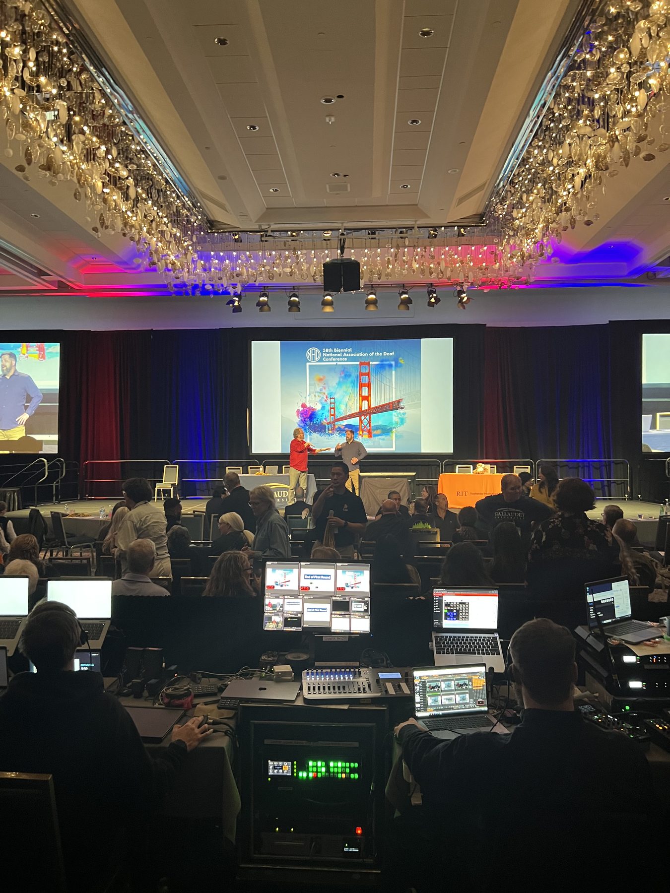 The main ballroom screen showing the 58th Biennial National Association of the Deaf Conference theme graphic, a colorful Golden Gate Bridge illustration, with the production booth and audience in the foreground.