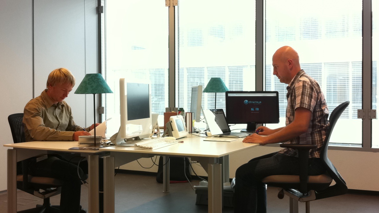 Inside DirectEye's new Ghent office in August 2011: two people working at facing desks along a wall of windows, with monitors, a laptop, a desktop video terminal and the DirectEye website open on the right-hand screen showing an NL / FR language choice.