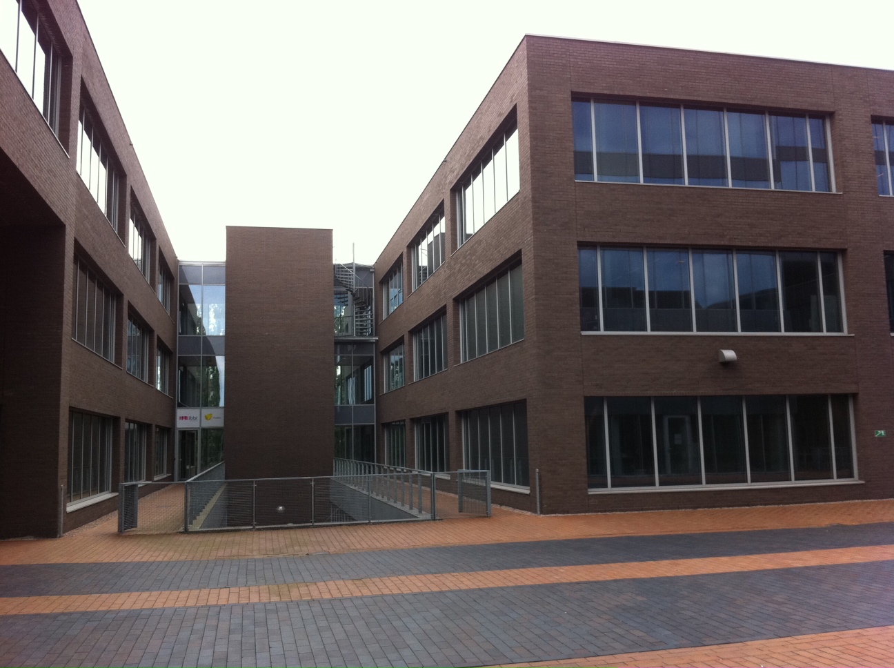 The courtyard of DirectEye's new Ghent office building in August 2011: two three-storey dark-brick office blocks with tall windows meeting at a glazed entrance, above a brick-paved plaza and a railed ramp.