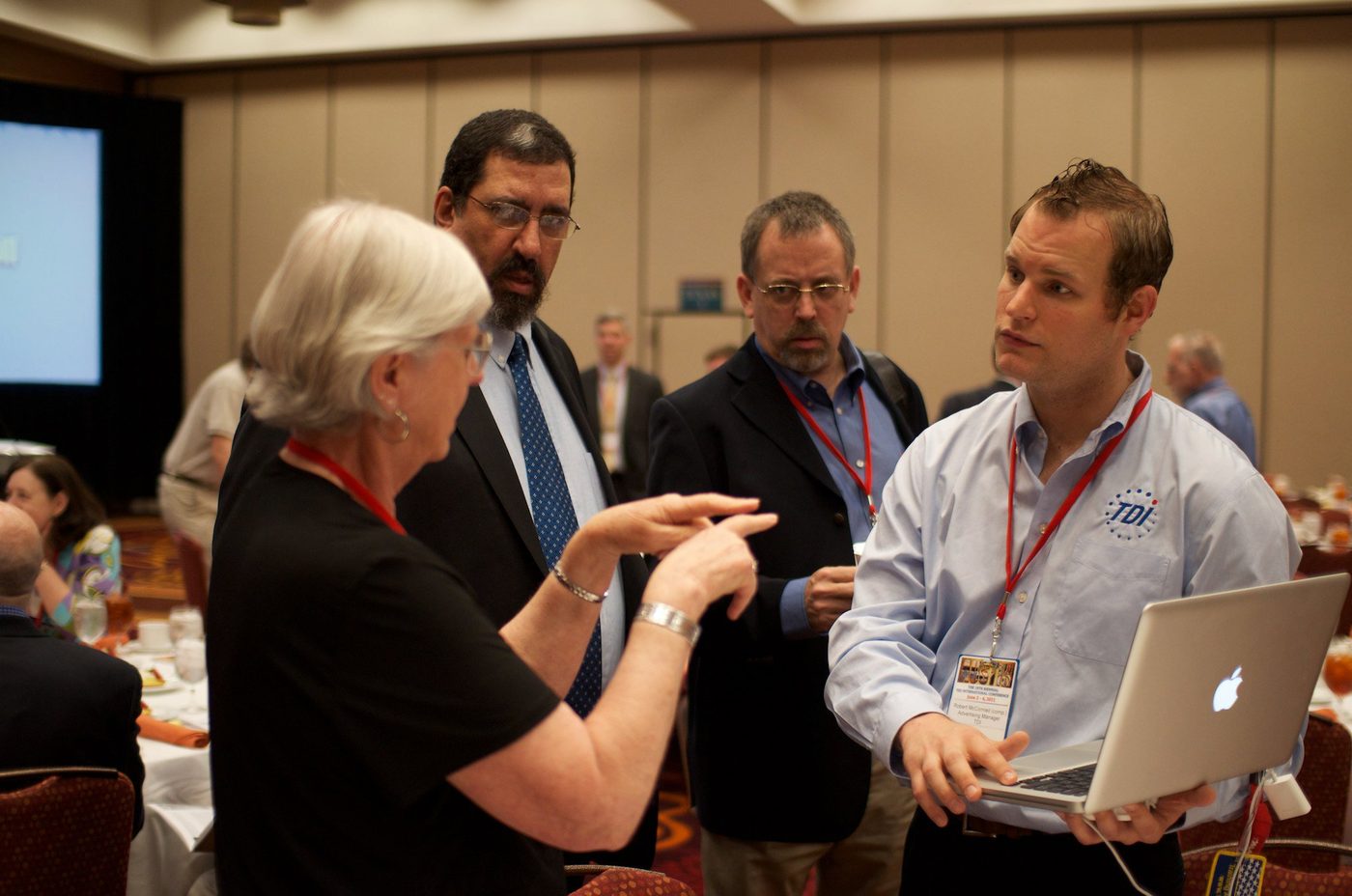 Four people conversing in sign language around an open laptop at the TDI conference; the person holding the laptop wears a TDI shirt and a conference badge.